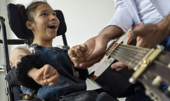 A young girl in a wheelchair smiles with excitement as an adult helps her engage with a guitar.