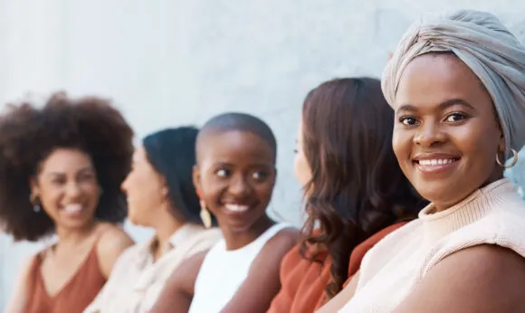 A group of women sit together along a wall, smiling and chatting, with one woman in the foreground looking directly at the camera.