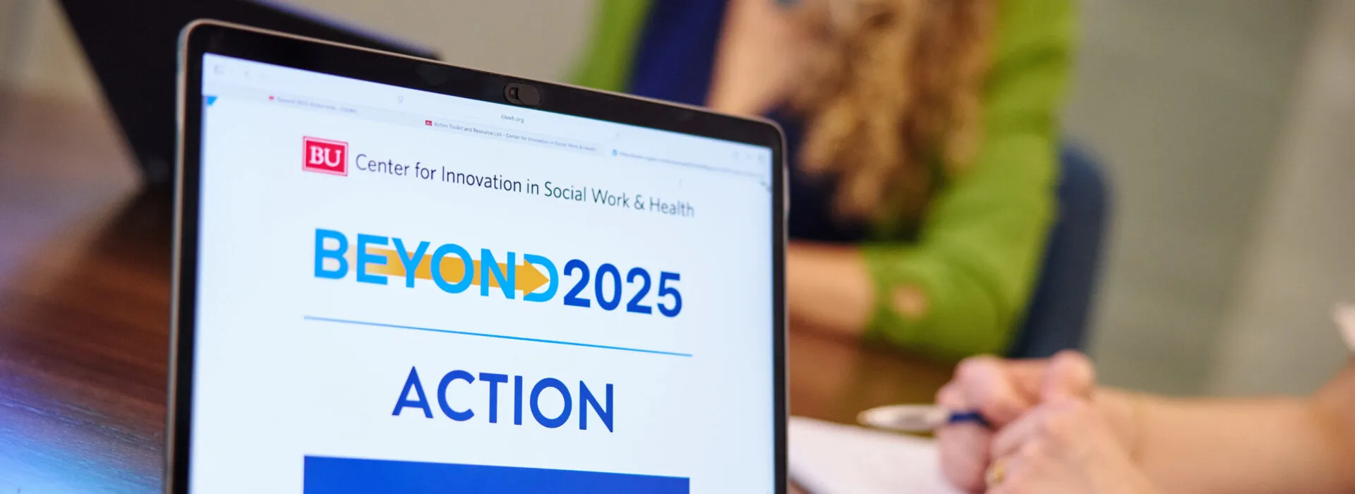 A laptop screen displays the Beyond 2025 Action website from the Center for Innovation in Social Work & Health, with people working and taking notes in the background.