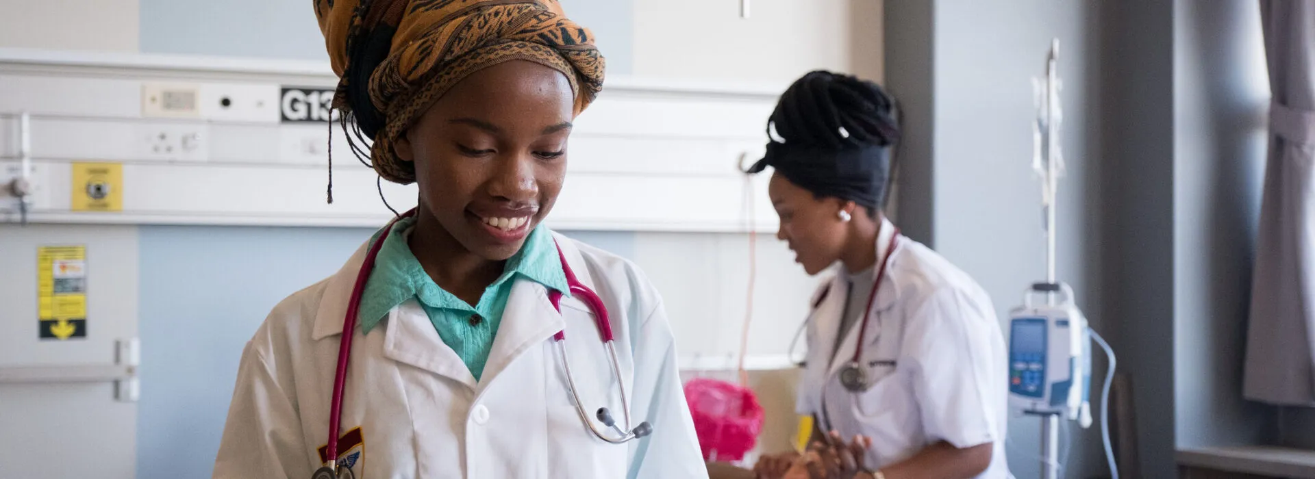 Two healthcare workers in a hospital room, one smiling while using a tablet and the other attending to medical equipment in the background.