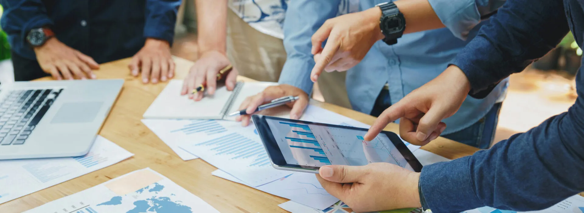 A group of people collaborate around a table covered with charts, papers, and a laptop, while one person uses a tablet to display data.
