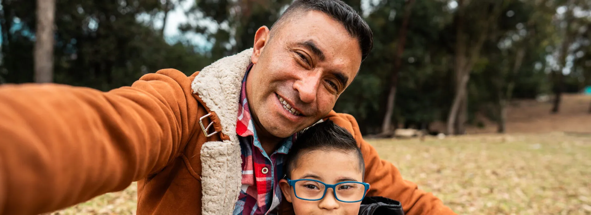 A father and son smile for a selfie while sitting together in a park on a cool day, surrounded by fallen leaves and trees.