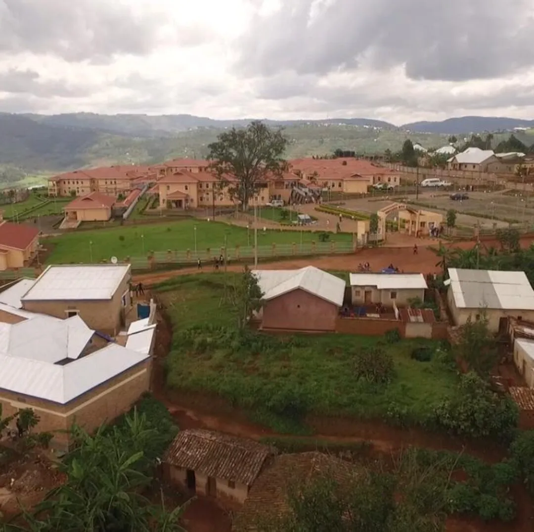 A zooming-out aerial view of a rural hillside community with small homes, greenery, and a larger institutional complex in the background, set against a backdrop of rolling hills and cloudy skies.