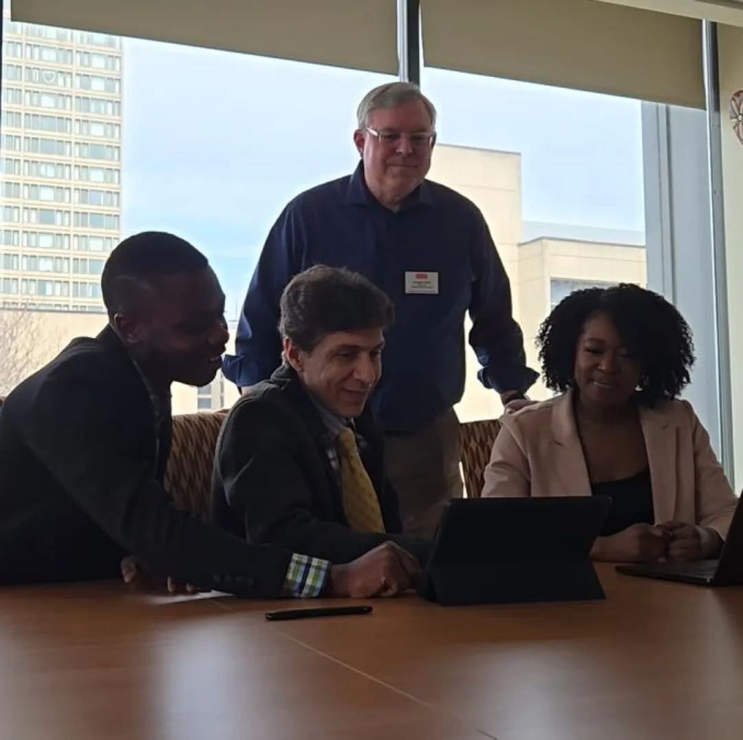 Four CISWH team members gather around a tablet at a conference table, collaborating during a meeting in a sunlit office with large windows and city buildings in the background.