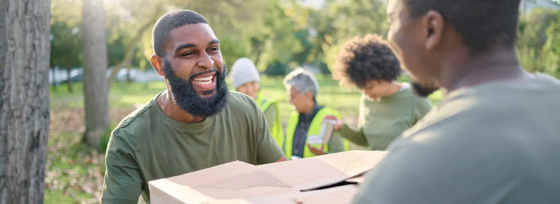 Smiling volunteers work together outdoors, passing a large box during a community service or donation event.